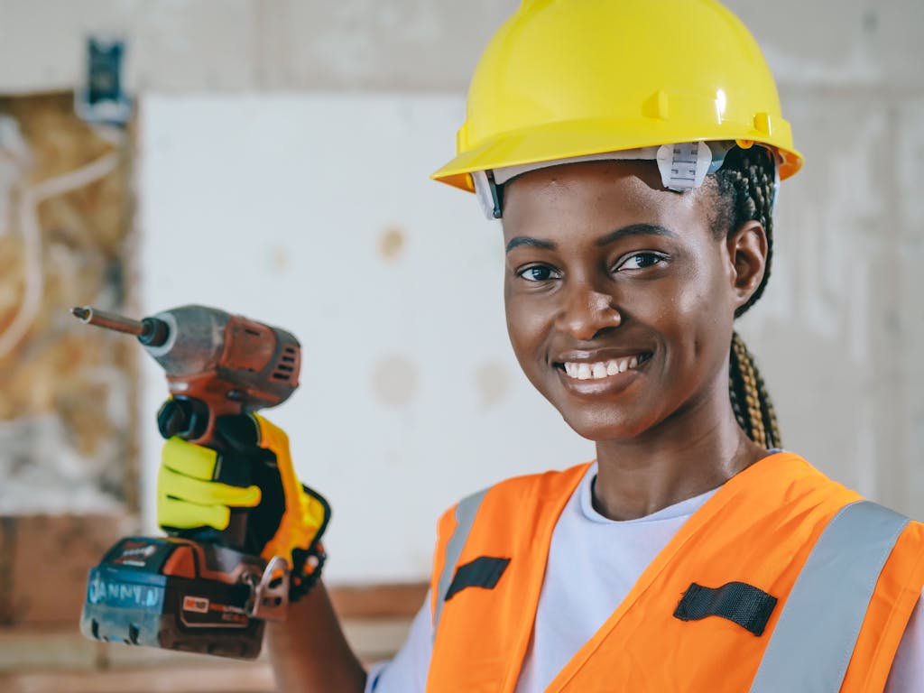 Confident female construction worker with hard hat and drill, smiling on site.